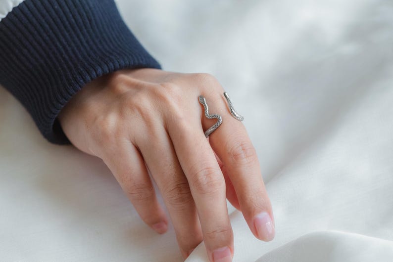 May include: A silver snake-shaped ring on a finger. The ring is made of two pieces of metal that are joined together in the shape of a snake.