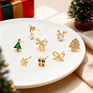 May include: A collection of gold-toned Christmas-themed stud earrings displayed on a white plate. The earrings feature various designs, including a Christmas tree, snowflake, bow, gift box, and reindeer. Some earrings are adorned with clear and green gemstones.