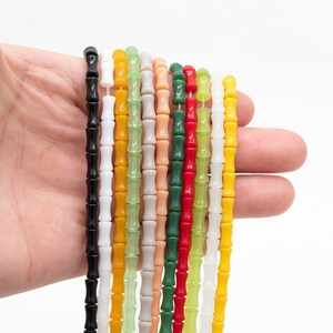 May include: A hand holding strands of colourful bamboo-shaped beads. The beads are in various colours, including black, white, yellow, green, red, and peach. The beads are strung together, creating a decorative element.