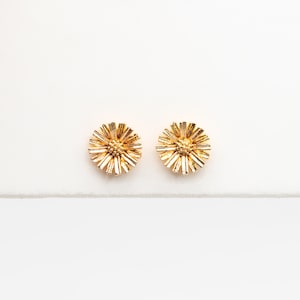 May include: A pair of gold-colored stud earrings. Each earring has a round, textured design resembling a flower with radiating petals. The earrings are set against a white background.