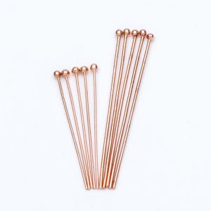 May include: A set of 12 rose gold head pins with rounded ends. The pins are arranged in two rows, with the longer pins in the back and the shorter pins in the front.
