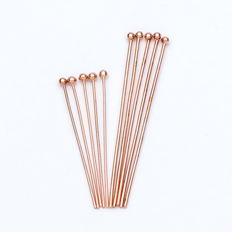 May include: A set of 12 rose gold head pins with rounded ends. The pins are arranged in two rows, with the longer pins in the back and the shorter pins in the front.