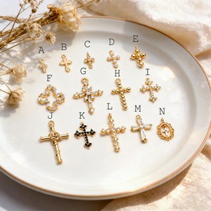 Pu&ograve; includere: Una collezione di ciondoli a forma di croce color oro con vari design, tra cui dettagli di perline, gemme e floreali. Le croci sono esposte su un piatto bianco, mostrando la loro intricata lavorazione artigianale e i diversi stili. Alcune croci hanno gemme chiare o nere.