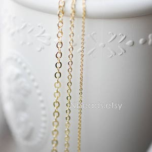 May include: Three gold chains of different lengths and thicknesses hanging from a white ceramic container. The chains are made of small, round links. The container has a decorative design.