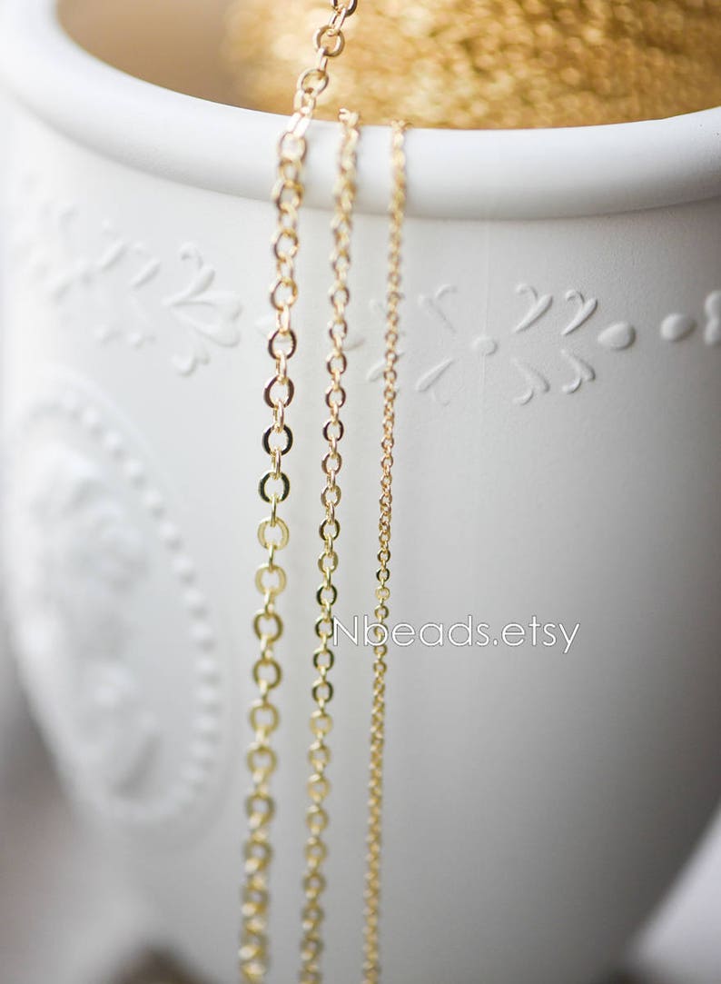 May include: Three gold chains of different lengths and thicknesses hanging from a white ceramic container. The chains are made of small, round links. The container has a decorative design.