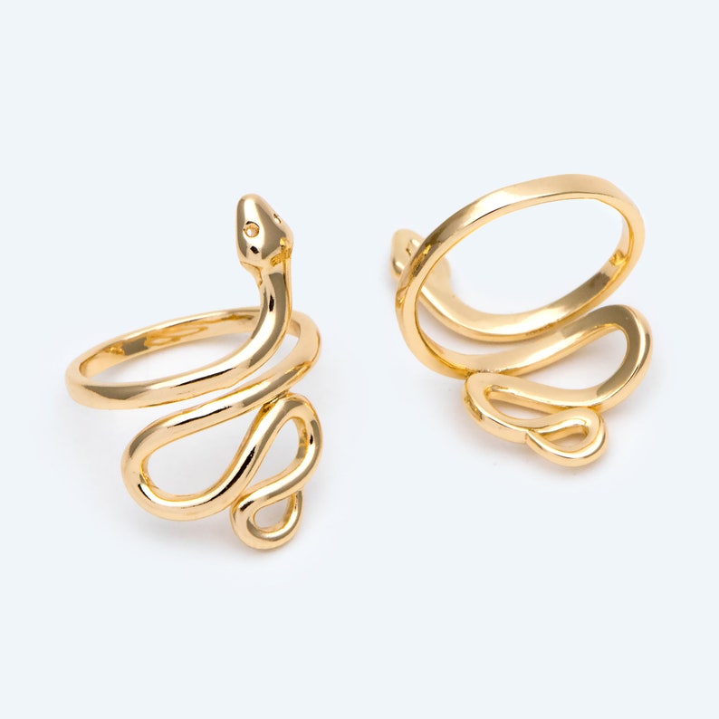 May include: Two gold-coloured snake rings. The rings feature a coiled snake design, with the snake's head and body wrapping around the finger. The rings are made of a shiny, metallic material and are set against a white background.