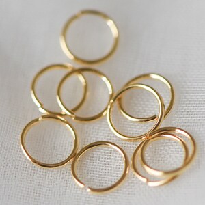 May include: A group of eight gold jump rings of various sizes, scattered on a white fabric background.