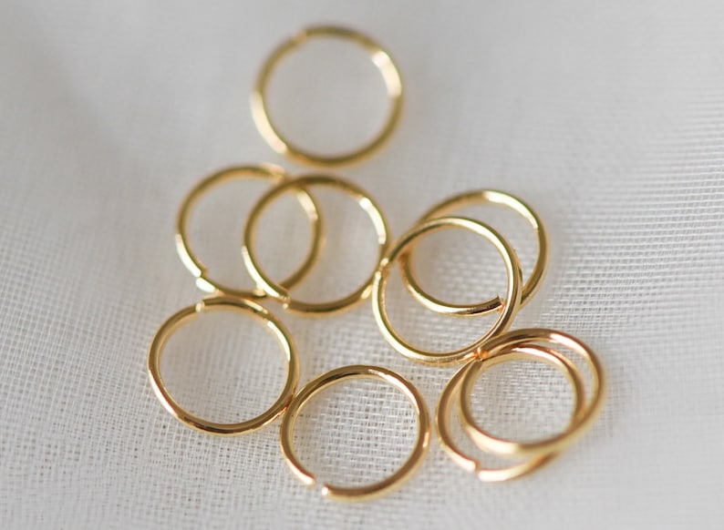 May include: A group of eight gold jump rings of various sizes, scattered on a white fabric background.