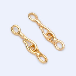 May include: Two gold-colored, decorative jewelry clasps. Each clasp features an oval loop at one end, a curved hook, and a decorative element. The clasps are designed to connect jewelry pieces.