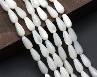 May include: Strands of pearly white teardrop-shaped beads. The beads are strung together, creating a flowing, elegant appearance. The beads have a smooth, glossy finish, reflecting light. The background is a dark wood and black surface.