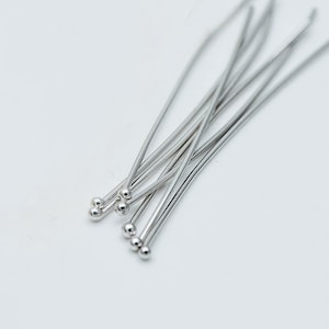 May include: A close-up of a group of silver head pins with round heads. The pins are arranged in a fan shape and are lying on a white surface.