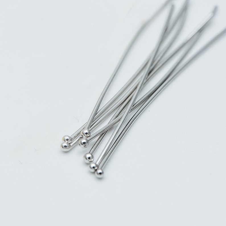 May include: A close-up of a group of silver head pins with round heads. The pins are arranged in a fan shape and are lying on a white surface.