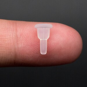 May include: A small, clear plastic bottle stopper with a flared top and a narrow, pointed bottom.