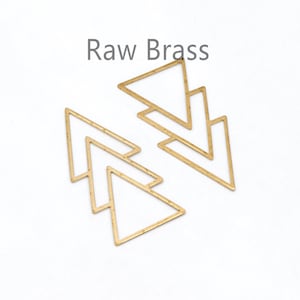 May include: Two raw brass arrow-shaped jewelry components. The open-frame design features a layered, geometric aesthetic. The text "Raw Brass" is printed in a dark gray font above the components. The components are set against a white background.