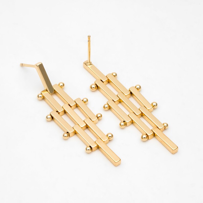 Puede incluir: Un par de pendientes geom&eacute;tricos de color dorado con un dise&ntilde;o largo y rectangular. Los pendientes est&aacute;n hechos de m&uacute;ltiples barras conectadas con peque&ntilde;as cuentas redondas en cada punto de conexi&oacute;n.