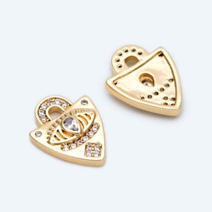 Puede incluir: Dos colgantes triangulares dorados con un lazo en la parte superior. Un colgante presenta una piedra transparente central en forma de lágrima rodeada de piedras transparentes más pequeñas. El otro colgante tiene un diseño similar. Probablemente sean componentes de joyería.