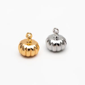 Könnte beinhalten: Zwei Kürbis-Anhänger, einer in Gold und einer in Silber, mit einer Öse oben zum Anbringen an Schmuck oder andere Bastelarbeiten.