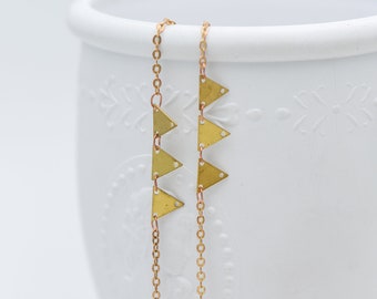 May include: A raw brass chain featuring small, flat, triangular metal charms with tiny holes at each point. The delicate gold-toned chain links are connected to these geometric shapes, which are arranged in a vertical line pattern.