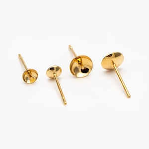 Op de afbeelding: Drie gouden oorbelpennen met ronde, platte toppen. De pennen zijn in verschillende maten.
