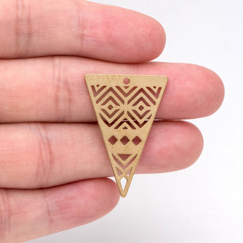 May include: A brass-colored, triangular pendant with a geometric cut-out design. The pendant features a series of diamond and triangle shapes, with a small hole at the top for stringing. This jewelry component is ideal for crafting earrings or necklaces.
