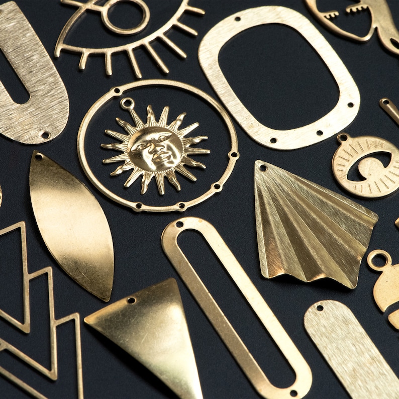 May include: A collection of gold-toned metal charms with various shapes and designs, including a sun, an eye, a geometric shape, and a leaf. The charms are arranged on a black background.