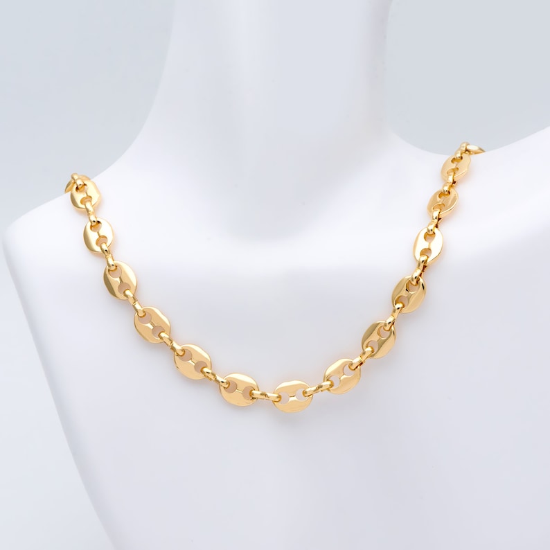 May include: A gold-colored necklace with a chain of flat, oval-shaped links. The necklace is displayed on a white mannequin bust. The links are evenly spaced and create a continuous, elegant design. The necklace has a bright, polished finish.