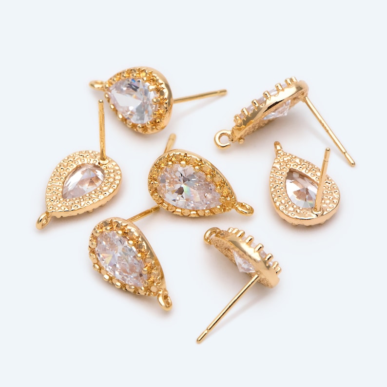 May include: Six gold teardrop-shaped earring findings with clear crystal stones. Each finding has a loop at the top and a post on the back.