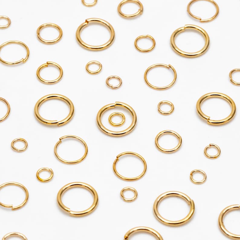 May include: A collection of gold jump rings of various sizes, arranged on a white background.