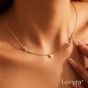 Op de afbeelding: Een goudkleurige ketting met de namen "Ayla" en "Sofia" in cursief schrift. Een kleine hartvormige bedel is gecentreerd tussen de namen. De ketting wordt op de borst van een persoon getoond.