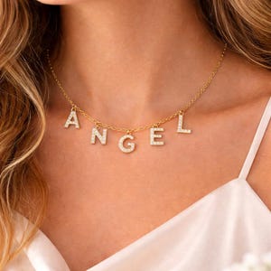 Peut inclure: Un collier doré avec le mot "ANGEL" épelé en lettres individuelles scintillantes incrustées de cristaux. Le collier est porté autour du cou, les lettres pendantes sous la chaîne. L'arrière-plan est la poitrine d'une femme et un haut blanc.