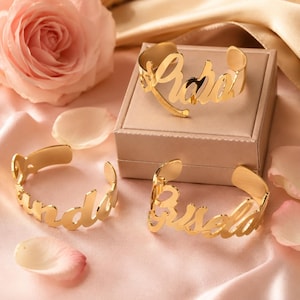 Könnte beinhalten: Goldfarbene Namensarmreifen, jeweils mit einem anderen Namen in Schreibschrift. Ein Armreif befindet sich auf einer Schachtel, zwei weitere auf einer rosa Oberfläche mit einer Rose und Blütenblättern.
