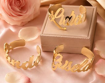 May include: Gold-colored name cuff bracelets, each with a different name in a cursive font. One bracelet is on a box, and two others are on a pink surface with a rose and petals.