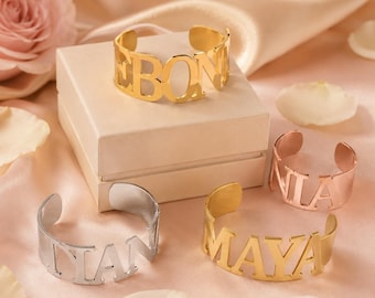 May include: Three open-cuff bracelets made of metal, featuring cut-out lettering. One bracelet is gold-toned with the name EBONI, one is rose gold-toned with the name NIA, and one is silver-toned with the name TIAN. A fourth gold-toned bracelet reads MAYA.