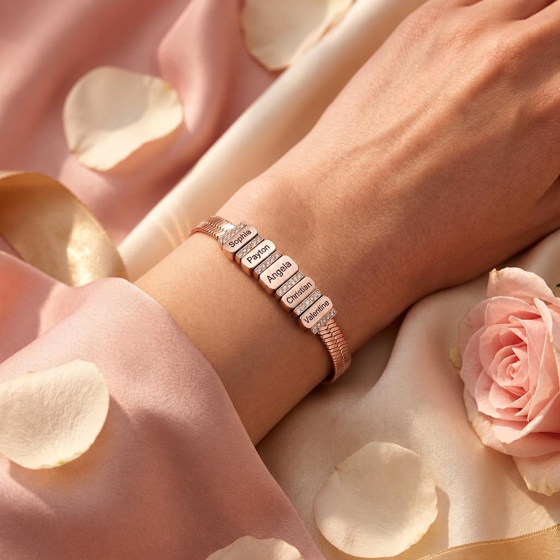 May include: A rose gold-toned bracelet featuring a flexible herringbone chain and five rectangular links. Each link is engraved with a name: Sophia, Payton, Angela, Christian, and Valentine. Small clear stones are set between each link.