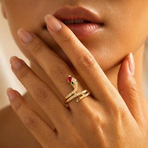 Può includere: Un anello a forma di serpente dorato con una testa di gemma rossa e verde e gemme trasparenti lungo il corpo. L'anello è su un dito, con la testa del serpente rivolta verso l'alto.