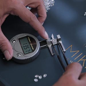K&ouml;nnte beinhalten: Ein digitaler Messschieber misst einen Diamanten, mit einer Anzeige von 13,57. Der Diamant wird mit einer Pinzette gehalten. Mehrere lose Diamanten sind auf einer dunkelblauen Oberfl&auml;che verstreut. Das Wort "MIKA" ist in goldfarbenen Buchstaben zu sehen.