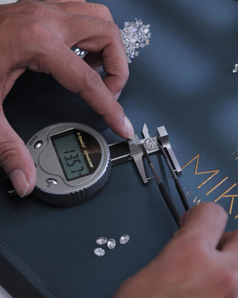 K&ouml;nnte beinhalten: Ein digitaler Messschieber misst einen Diamanten, mit einer Anzeige von 13,57. Der Diamant wird mit einer Pinzette gehalten. Mehrere lose Diamanten sind auf einer dunkelblauen Oberfl&auml;che verstreut. Das Wort "MIKA" ist in goldfarbenen Buchstaben zu sehen.