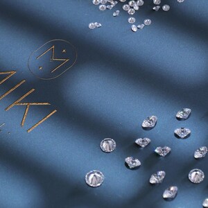 K&ouml;nnte beinhalten: Diamanten verstreut auf einem dunkelblauen Stoff. Das Bild zeigt ein Logo mit dem Wort "MIKI" in goldener Schrift. Die Diamanten sind in verschiedenen Gr&ouml;&szlig;en und Formen und reflektieren das Licht.