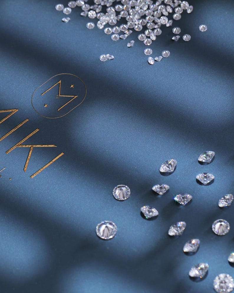 K&ouml;nnte beinhalten: Diamanten verstreut auf einem dunkelblauen Stoff. Das Bild zeigt ein Logo mit dem Wort "MIKI" in goldener Schrift. Die Diamanten sind in verschiedenen Gr&ouml;&szlig;en und Formen und reflektieren das Licht.