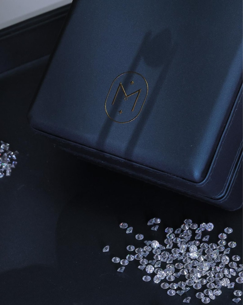 K&ouml;nnte beinhalten: Eine marineblaue Schmuckschatulle mit einem goldfarbenen "M"-Logo und verstreuten Diamanten auf einer schwarzen Oberfl&auml;che. Die Schatulle ist rechteckig und scheint aus Leder zu sein. Die Diamanten sind klein und funkelnd.