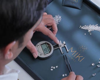 Pode incluir: Uma pessoa mede um diamante com um paquímetro digital em uma bandeja azul escura. A bandeja tem o logotipo dourado "MIKI JEWEL" e uma pequena bolsa com o mesmo logotipo. Vários diamantes soltos estão espalhados pela bandeja.