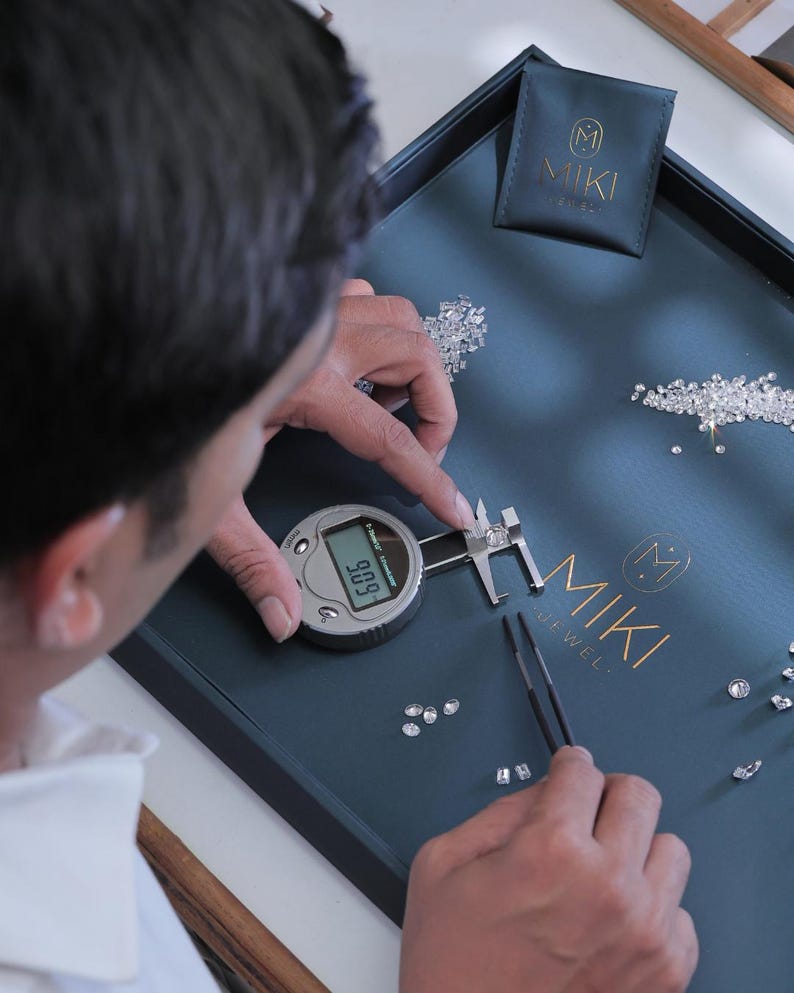 K&ouml;nnte beinhalten: Eine Person misst einen Diamanten mit einem digitalen Messschieber auf einem dunkelblauen Tablett. Das Tablett tr&auml;gt das goldene Logo "MIKI JEWEL" und einen kleinen Beutel mit demselben Logo. Mehrere lose Diamanten sind auf dem Tablett verstreut.
