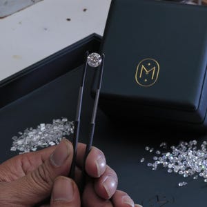 K&ouml;nnte beinhalten: Ein runder Diamant im Brillantschliff wird von einer Pinzette gehalten. Der Diamant ist vor einem dunklen Hintergrund mit verstreuten Diamanten und einer dunkelgr&uuml;nen Schatulle mit goldenem Emblem platziert. Das Bild zeigt die Klarheit und das Funkeln des Diamanten.