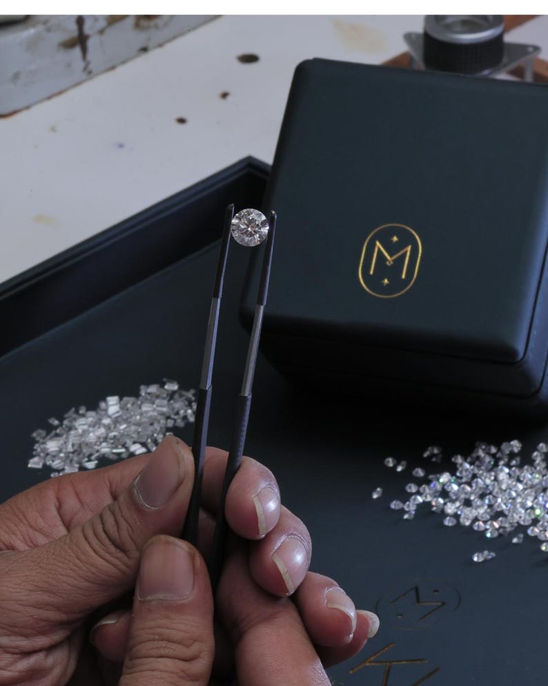 K&ouml;nnte beinhalten: Ein runder Diamant im Brillantschliff wird von einer Pinzette gehalten. Der Diamant ist vor einem dunklen Hintergrund mit verstreuten Diamanten und einer dunkelgr&uuml;nen Schatulle mit goldenem Emblem platziert. Das Bild zeigt die Klarheit und das Funkeln des Diamanten.