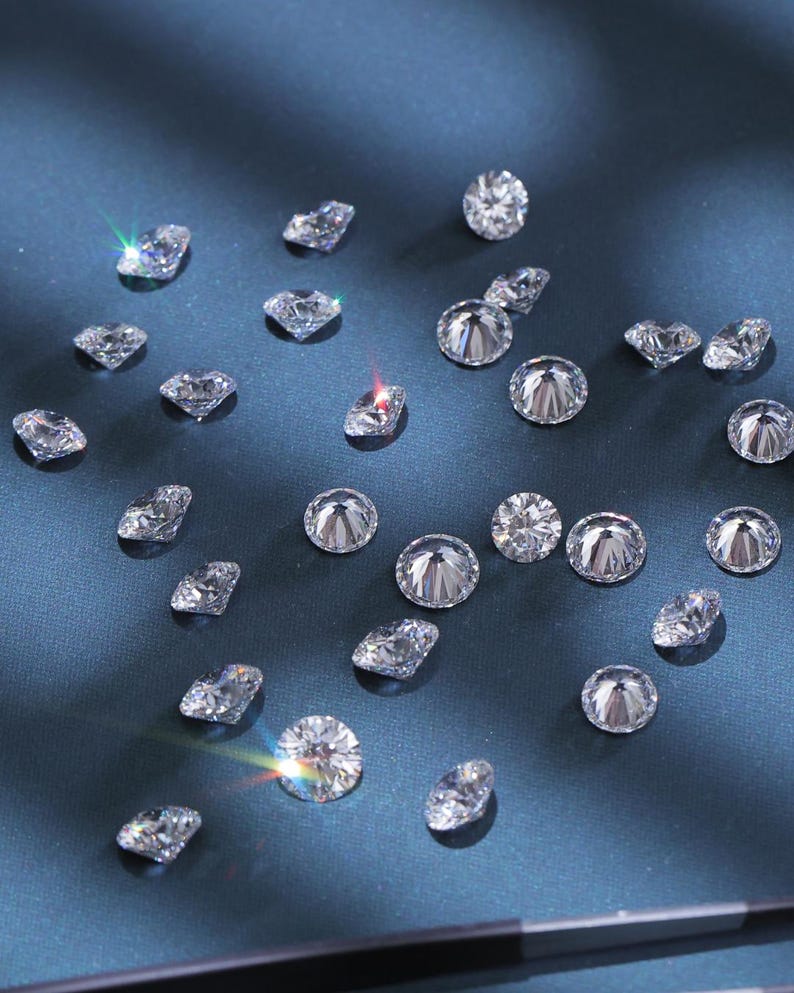 K&ouml;nnte beinhalten: Verstreute, klare Edelsteine ​​verschiedener Formen und Gr&ouml;&szlig;en, darunter runde und facettierte Schliffe, funkeln vor einem dunkelblauen Hintergrund. Die Edelsteine ​​reflektieren das Licht und erzeugen ein brillantes Funkeln. Das Bild zeigt eine Sammlung loser Diamanten.