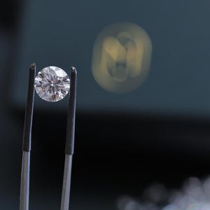 K&ouml;nnte beinhalten: Ein runder Diamant im Brillantschliff, gehalten von einer Pinzette. Der Diamant funkelt mit zahlreichen Facetten, die das Licht reflektieren. Der Hintergrund ist ein verschwommenes Dunkelblaugr&uuml;n mit einer weichen, unscharfen Lichtquelle.