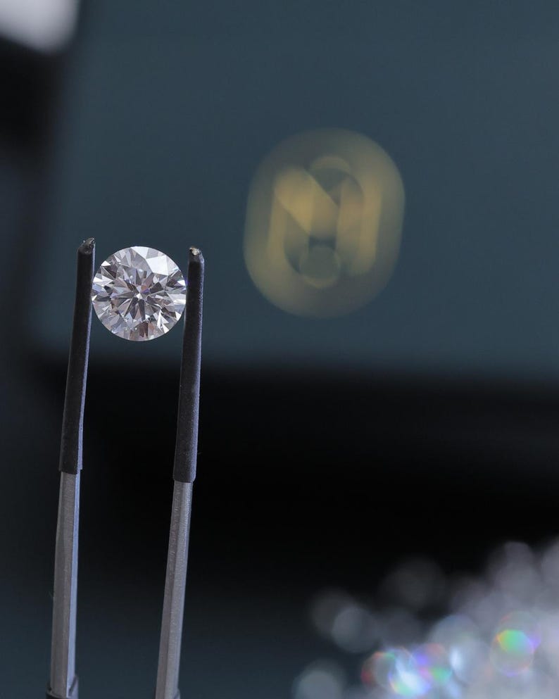 K&ouml;nnte beinhalten: Ein runder Diamant im Brillantschliff, gehalten von einer Pinzette. Der Diamant funkelt mit zahlreichen Facetten, die das Licht reflektieren. Der Hintergrund ist ein verschwommenes Dunkelblaugr&uuml;n mit einer weichen, unscharfen Lichtquelle.