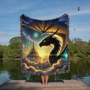 May include: A woven blanket featuring a fantasy dragon design. The artwork depicts a black dragon with green eyes, a castle, and a celestial background with clouds and stars. The blanket has a fringed edge.