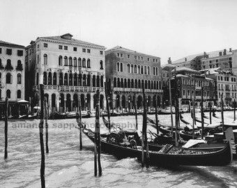 May include: Black and white photograph of the Grand Canal in Venice, Italy. Several gondolas are docked along the water's edge, with buildings lining the canal. Wooden poles are in the water. The sky is overcast.