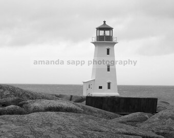 May include: A white lighthouse with a black roof stands on a rocky shore. The lighthouse is tall and slender, with a small window on each side. The sky is overcast and the water is choppy.